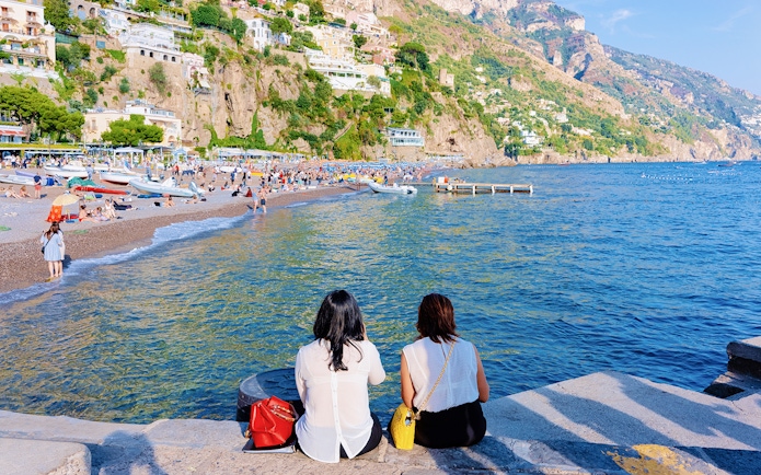 Two people sitting by the sea in Positano, with a view of the beach and hillside houses.