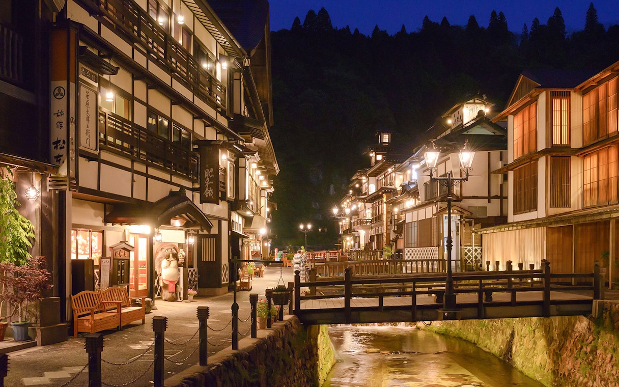 Traditional Japanese street in Ginzan Onsen, Tohoku, illuminated at night, showcasing wooden buildings and a bridge.