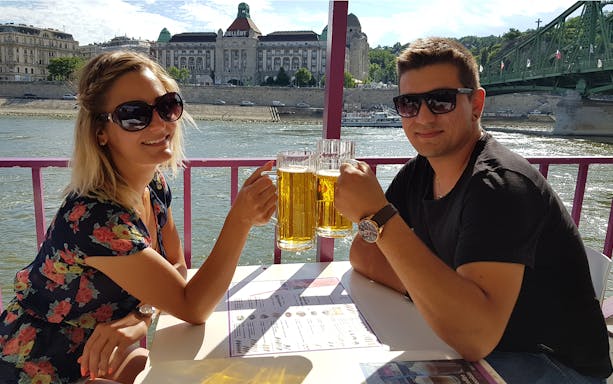 Couple enjoying drinks on a Budapest cruise with Gellért Hotel and Liberty Bridge in the background.