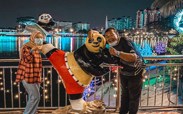 Visitors posing with panda statue at 99 Wonderland Park, colorful lights reflecting on water.