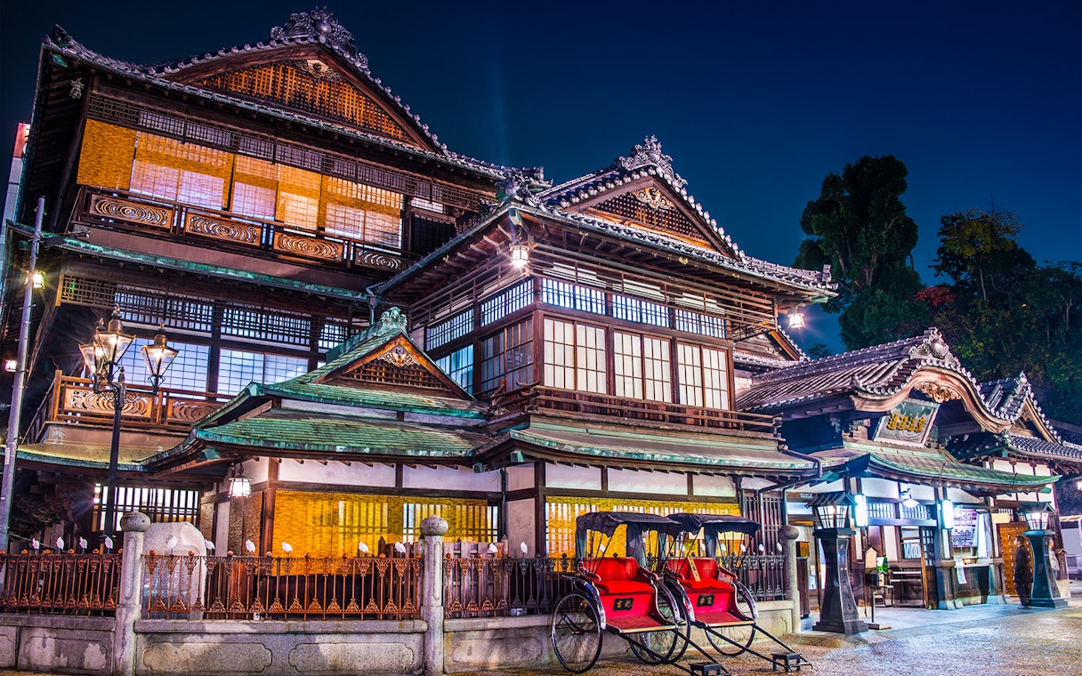 Historic Dogo Onsen building illuminated at night, Matsuyama, Japan, with rickshaws in front.