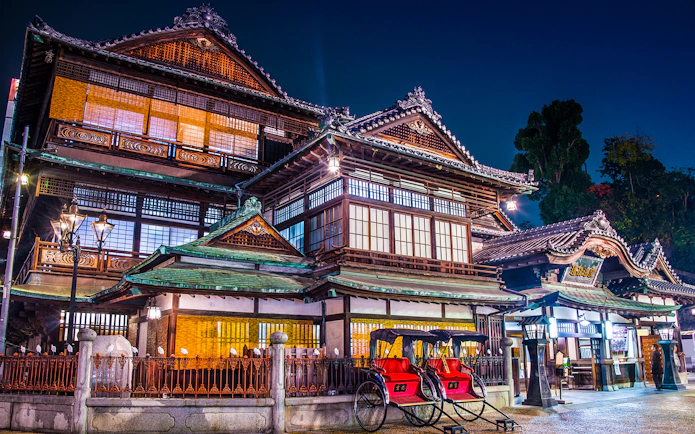 Historic Dogo Onsen building illuminated at night, Matsuyama, Japan, with rickshaws in front.