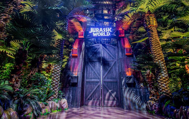 Entrance gate to Jurassic World: The Exhibition in London surrounded by lush greenery.