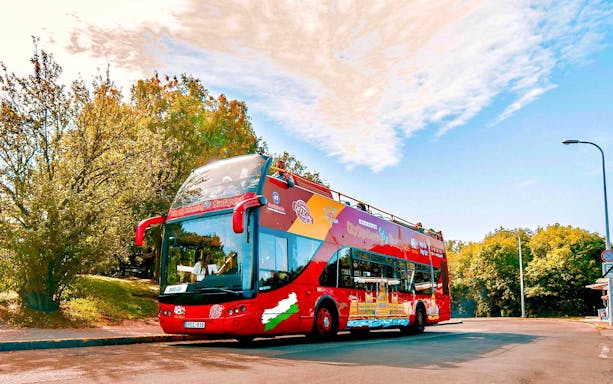 Red double-decker bus for Budapest hop-on hop-off tour parked near trees.