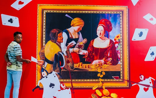 Man interacting with 3D art at Trick Eye Museum, featuring a classic painting and playing cards.