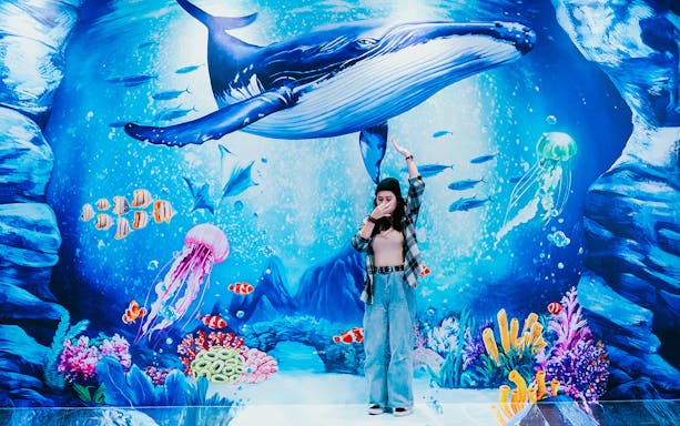 Visitor posing with underwater mural at Trick Eye Museum, featuring a whale and colorful sea life.
