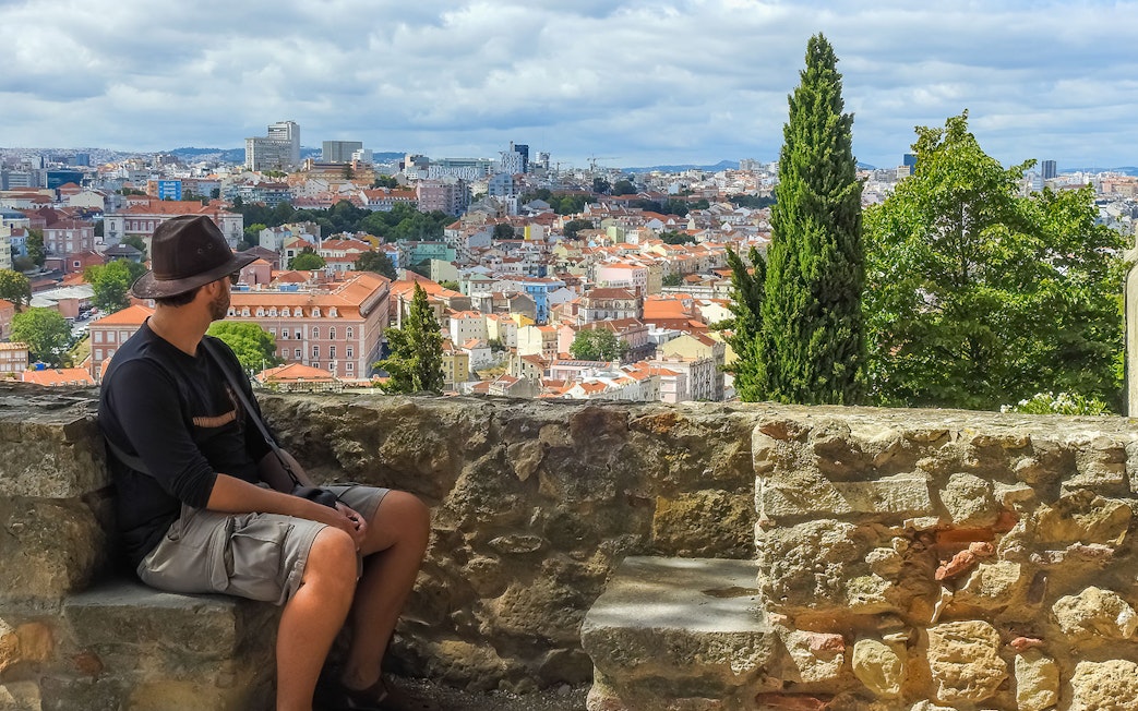 Man seated on St. George’s Castle wall overlooking Lisbon cityscape.