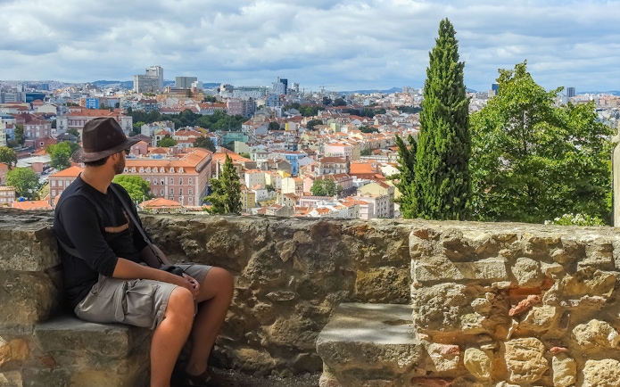Man seated on St. George’s Castle wall overlooking Lisbon cityscape.