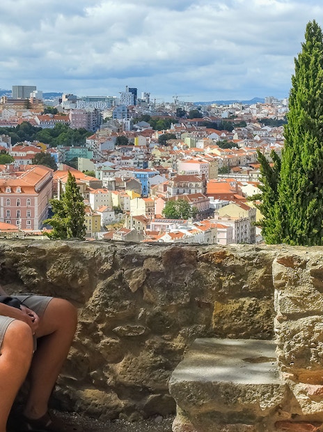 Man seated on St. George’s Castle wall overlooking Lisbon cityscape.