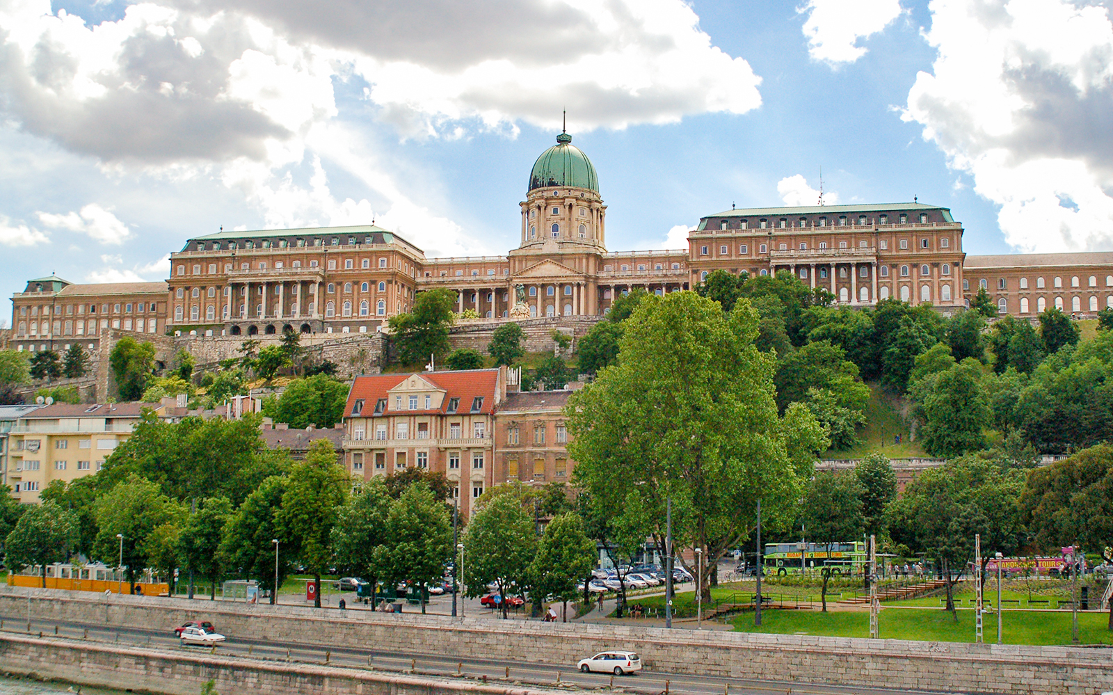 Buda Castle in Budapest viewed from the Danube River, surrounded by lush greenery.