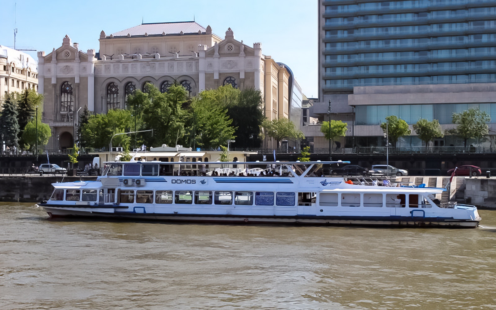 Danube River cruise boat with tourists and Budapest landmarks in view.