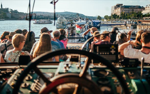 Danube River cruise with tourists seated, Budapest skyline in background.