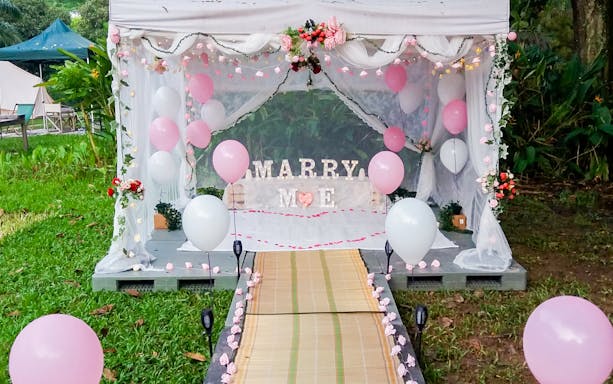 Glamping tent with "Marry Me" sign and pink balloons in a garden setting.