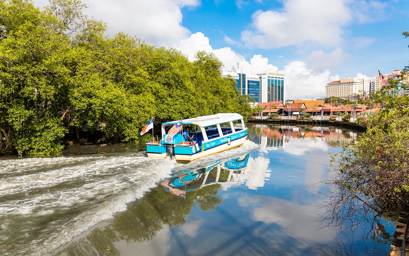 Boat cruising on Melaka River with cityscape and lush greenery.