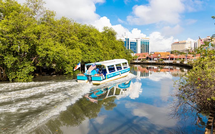 Boat cruising on Melaka River with cityscape and lush greenery.