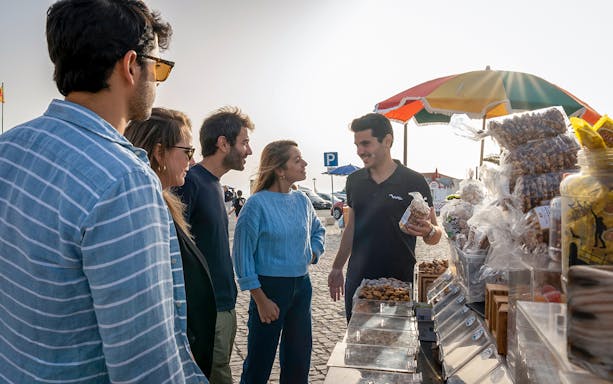 Tourists interacting with a vendor at a market stall during a guided tour from Lisbon to Porto, Nazaré, and Óbidos.