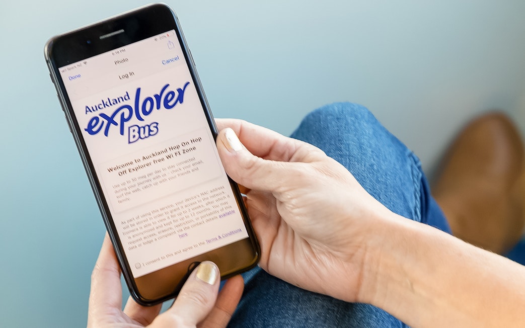 Person holding a phone displaying Auckland Explorer Bus hop-on-hop-off tour information.