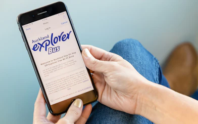 Person holding a phone displaying Auckland Explorer Bus hop-on-hop-off tour information.
