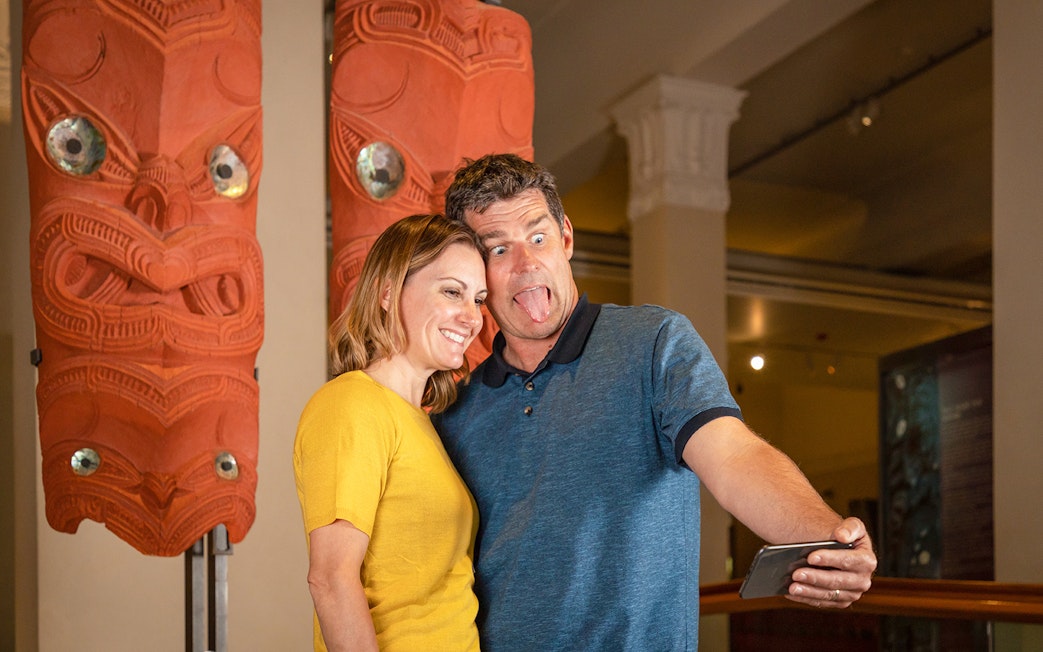 Couple taking selfie with Māori carvings at Auckland museum on hop-on-hop-off tour.