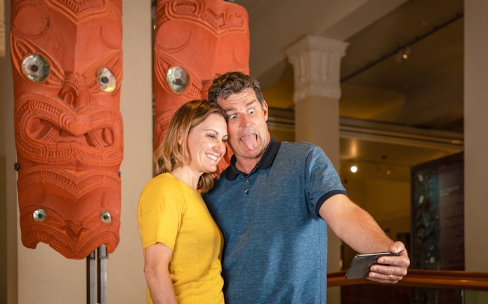 Couple taking selfie with Māori carvings at Auckland museum on hop-on-hop-off tour.