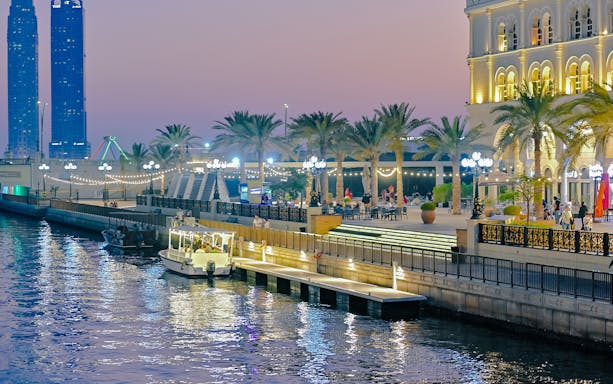 Private boat docked at Khalid Lagoon with illuminated promenade and palm trees in the evening.