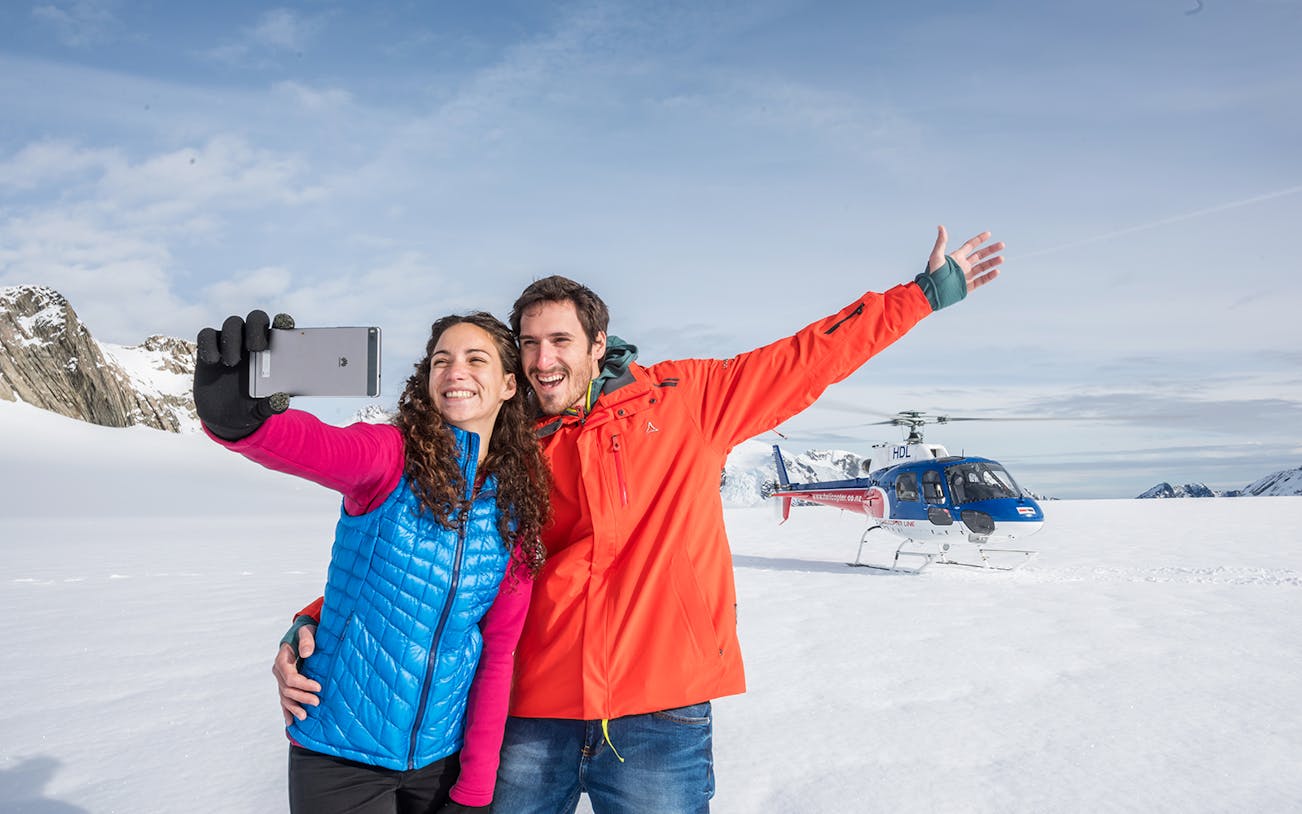 Couple taking selfie on snowfield with helicopter during Neve Discoverer Tour, Franz Josef.