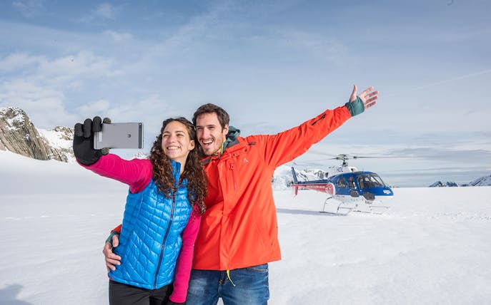 Couple taking selfie on snowfield with helicopter during Neve Discoverer Tour, Franz Josef.