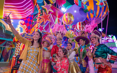 Carnival performers and visitors at Carnival Magic Park, vibrant costumes and lights.