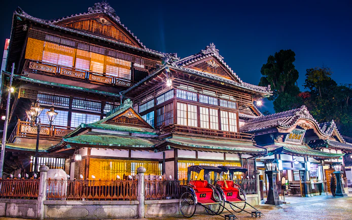 Dogo Onsen Honkan in Matsuyama, Japan, illuminated at night with traditional rickshaws in front.