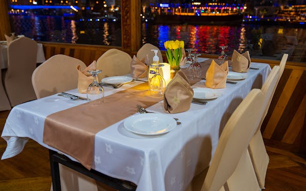 Dinner table setup on a Dhow Cruise, Dubai Creek, with city lights reflecting on water.