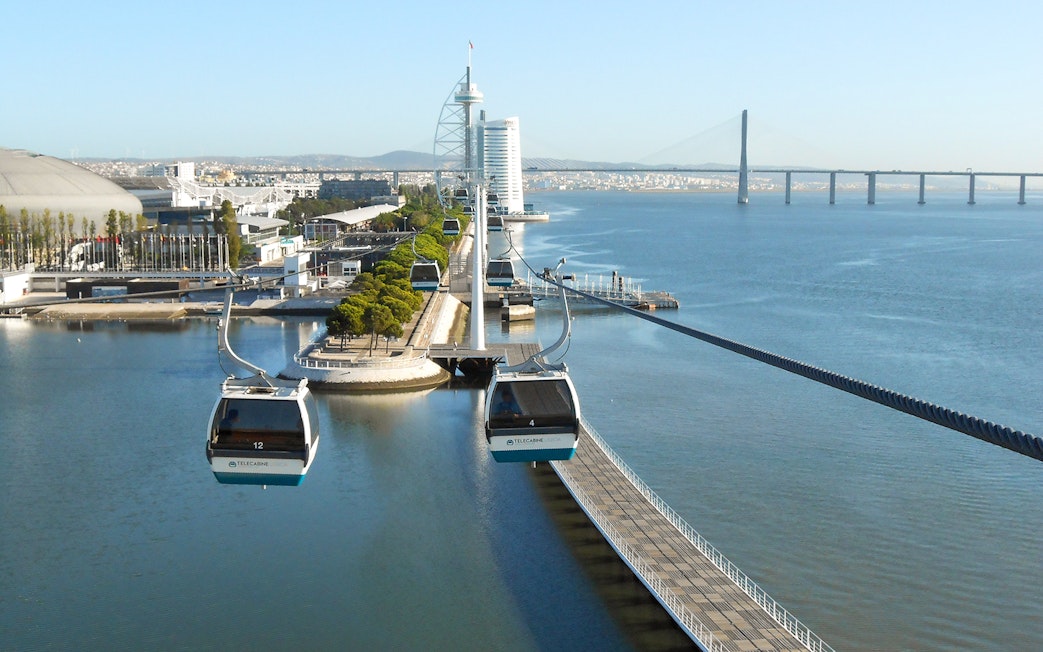 Telecabine cable cars over Tagus River in Lisbon with cityscape and bridge in view.
