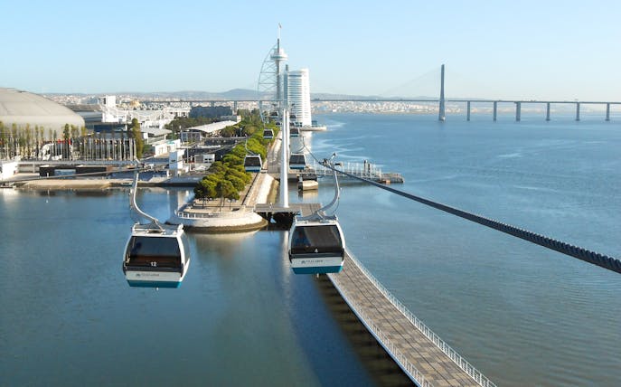 Telecabine cable cars over Tagus River in Lisbon with cityscape and bridge in view.