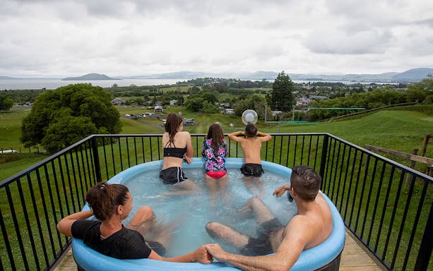 Family relaxing in a hot tub overlooking Zorb Rotorua landscape, New Zealand.