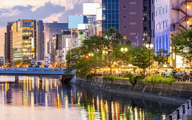 Cityscape of Fukuoka at dusk with illuminated buildings and riverside food stalls, Japan.