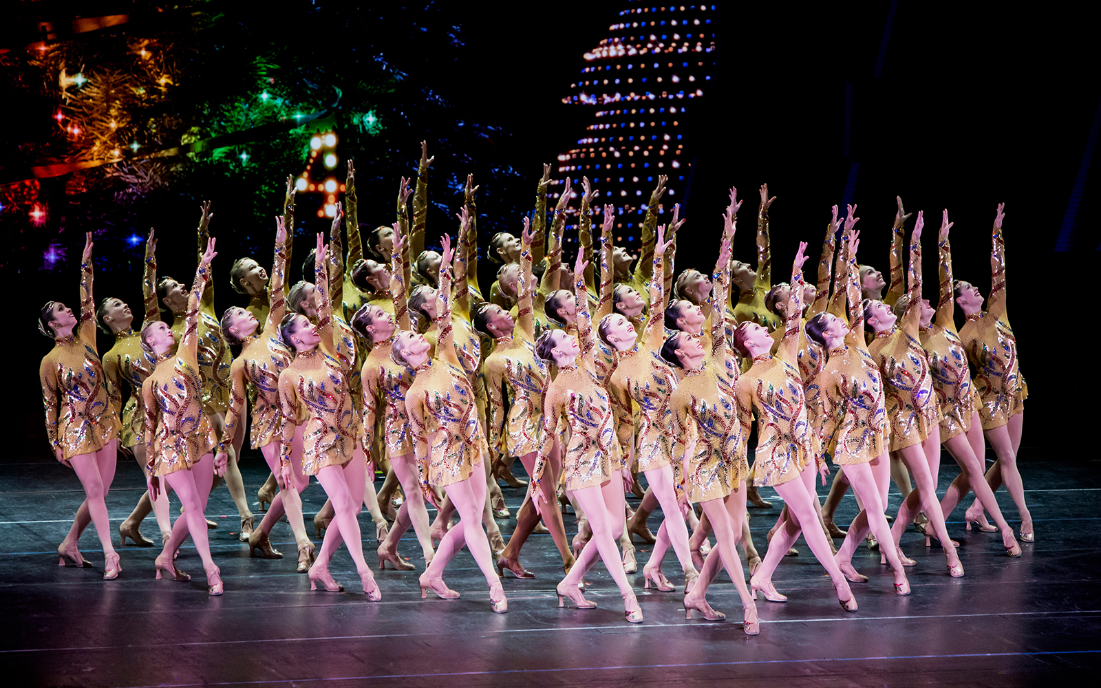 The rockettes in a formation and sparkly dresses, their legs crossed, left arm outstretched overhead.