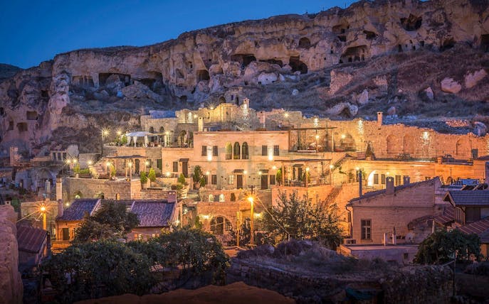 Cave hotel illuminated at dusk in Cappadocia, Turkey, with rock formations in the background.