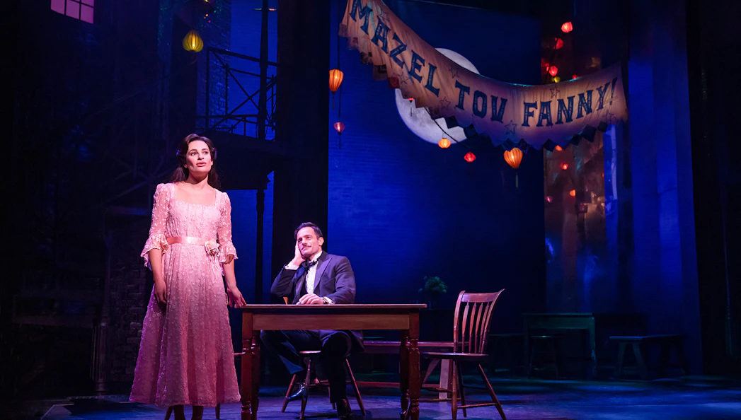 Stage scene from "Funny Girl" with a woman in a pink dress and a man seated at a table under a "Mazel Tov Fanny" banner.
