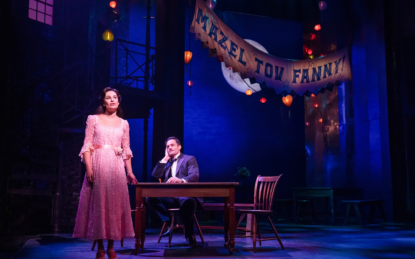 Stage scene from "Funny Girl" with a woman in a pink dress and a man seated at a table under a "Mazel Tov Fanny" banner.
