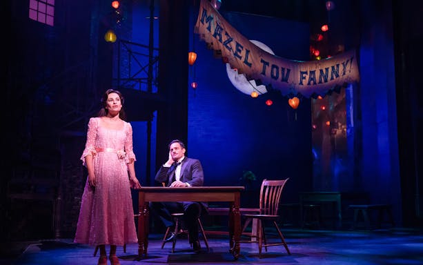 Stage scene from "Funny Girl" with a woman in a pink dress and a man seated at a table under a "Mazel Tov Fanny" banner.