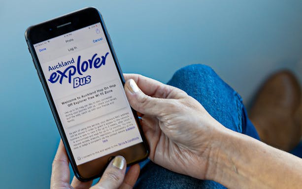 A person holding a phone displaying Auckland Explorer Bus information.