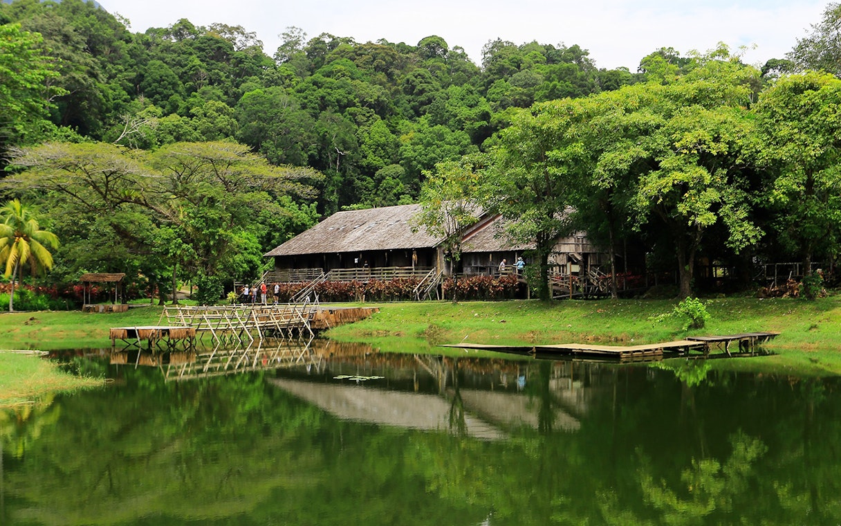 Traditional longhouse by a pond at Sarawak Cultural Village, surrounded by lush greenery.
