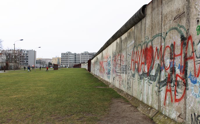 Berlin Wall with graffiti, tourists walking nearby on a guided tour.