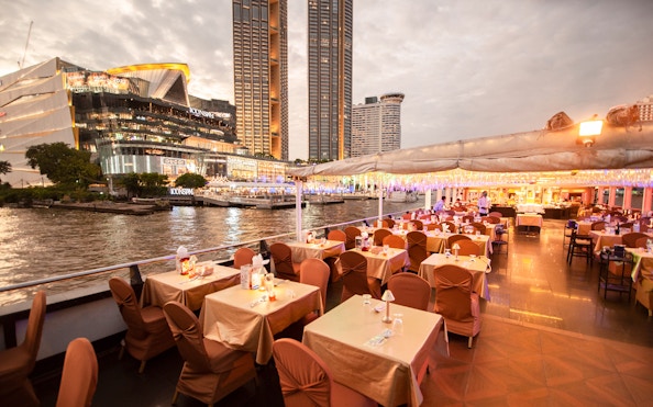 Chao Phraya dinner cruise with tables set for dining, view of Iconsiam and riverfront in Bangkok.