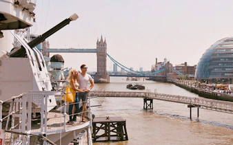 Couple on HMS Belfast deck with Tower Bridge in London background.