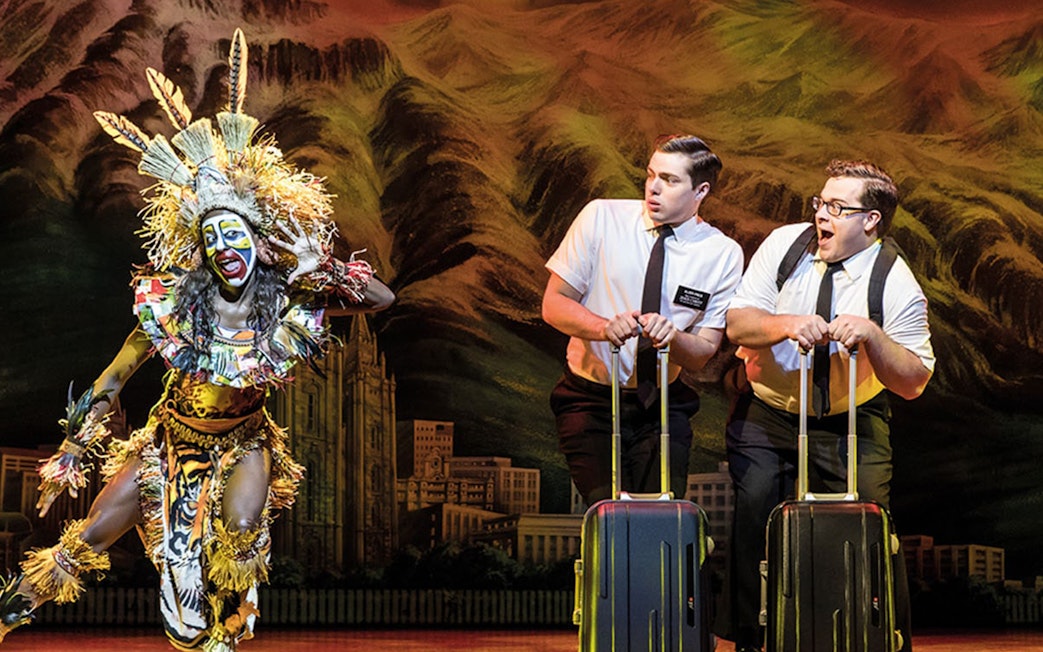 Performers in "The Book of Mormon" musical scene with vibrant costumes and props.
