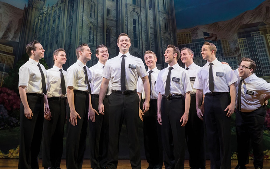 Cast of "The Book of Mormon" performing on stage with a backdrop of a cityscape.