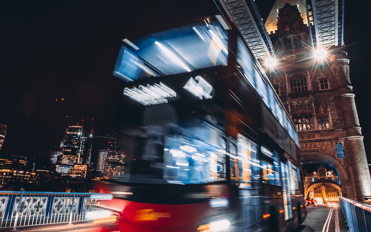 Double-decker bus crossing Tower Bridge at night, London skyline in background.