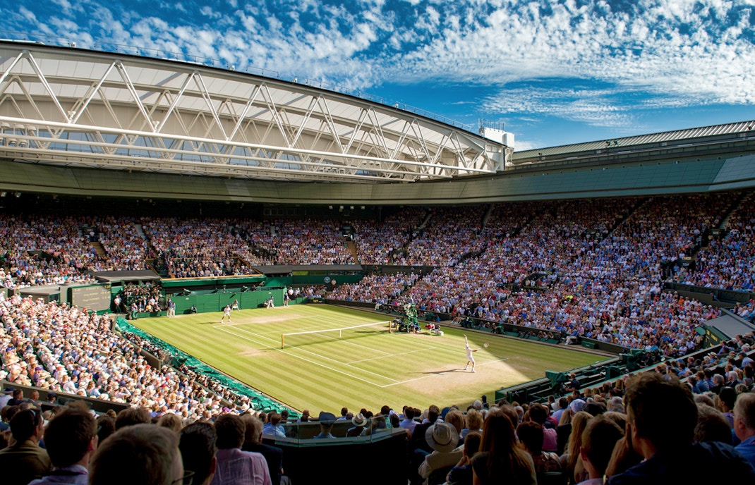 Wimbledon Centre Court during a tennis match, with a full crowd under a clear sky.