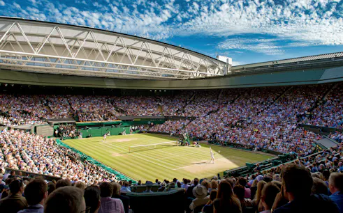 Wimbledon Centre Court during a tennis match, with a full crowd under a clear sky.