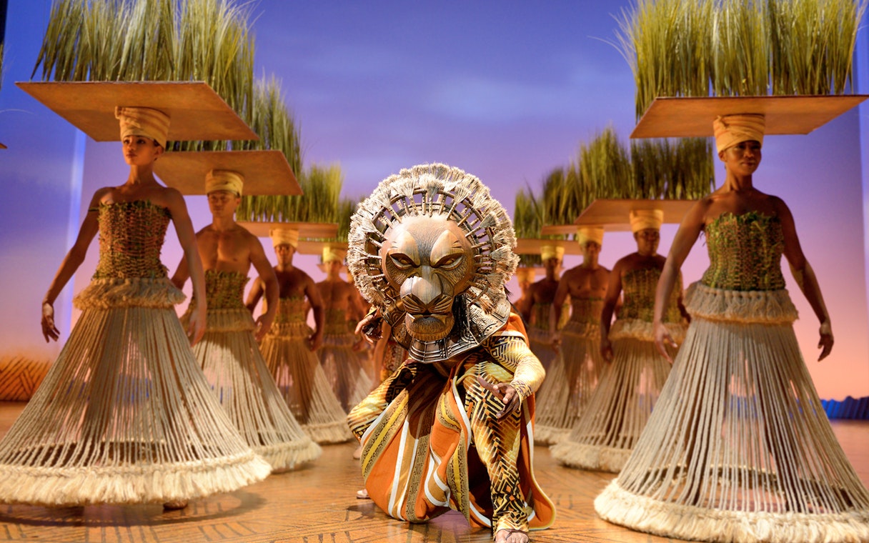 Lion King stage performance with actors in traditional costumes and lion mask.